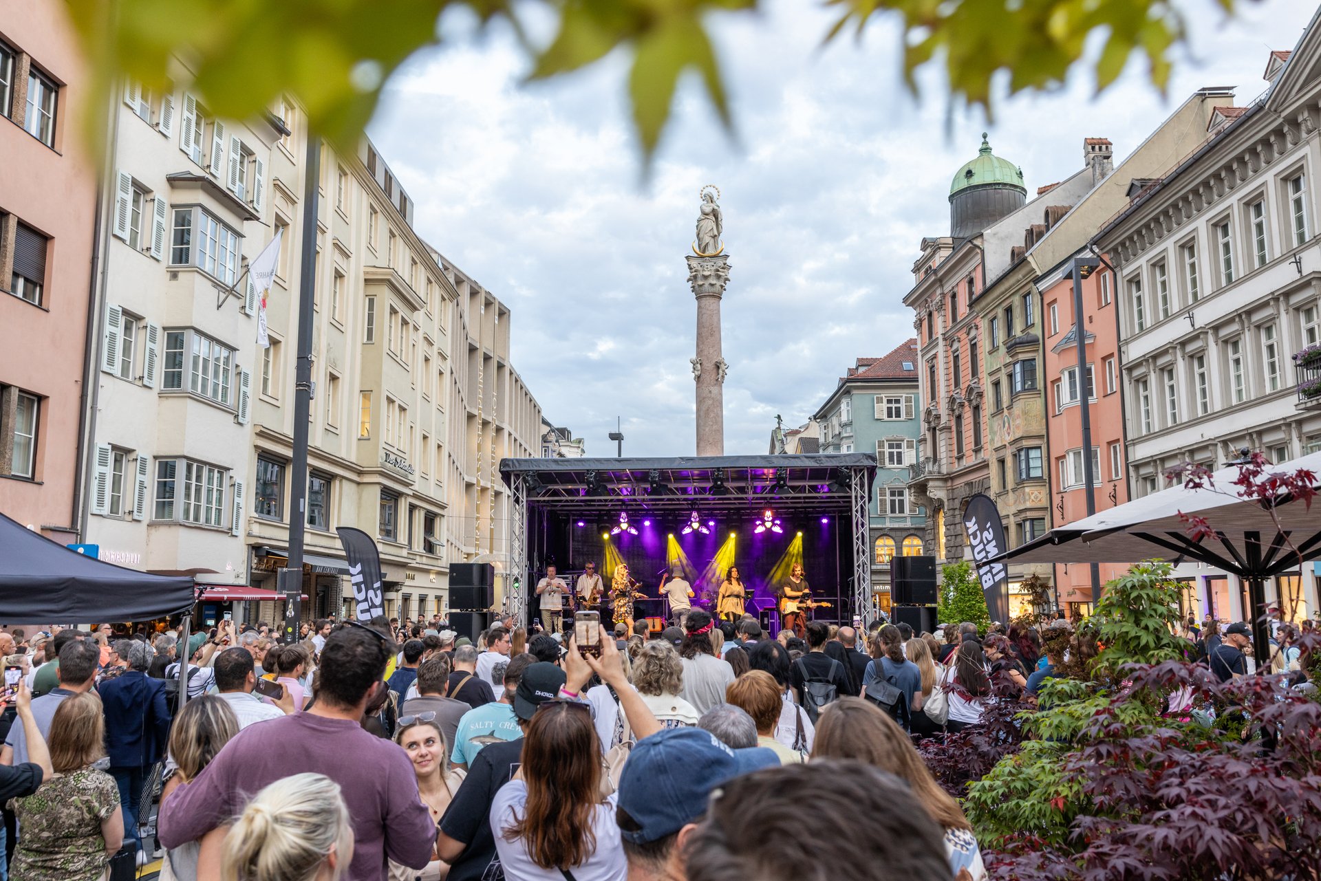Große Menschenmenge bei einer Open-Air-Bühne, Band spielt auf Bühne, in der Innsbrucker Maria-Theresien-Straße; vor historischer Häuserkulisse spielt eine Band auf einer beleuchteten Bühne, im Hintergrund ragt die Annasäule empor.